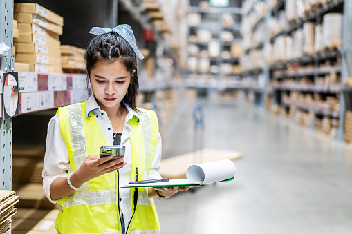 Young asian woman worker uses the phone for business contacts and holding clipboard to checking inventory in the warehouse
