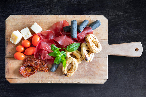 A chopping board with tomatoes, cheese, bread, bresaola, and herbs