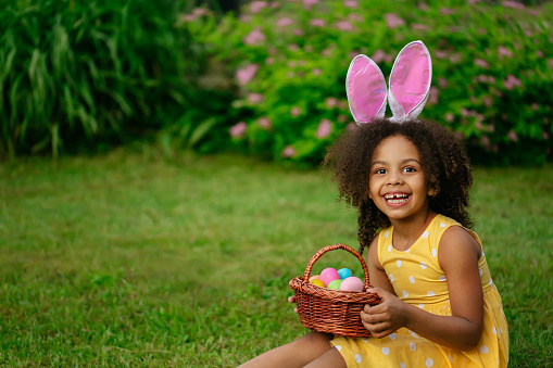 Little Black girl wear bunny ears and gathering Easter eggs on Easter egg hunt in garden. Child with busket full of colored eggs