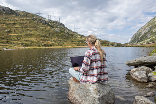 Young woman working remotely, sits on a rock surrounded by nature