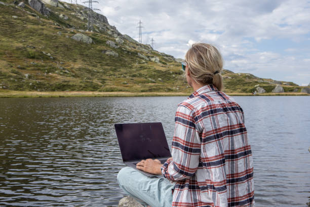 istock Woman sits on rock using laptop by the lake, remote working 1465524011