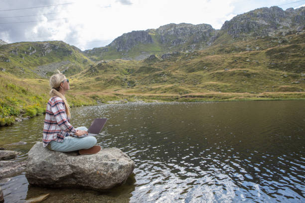 istock Woman sits on rock using laptop by the lake, remote working 1465525036