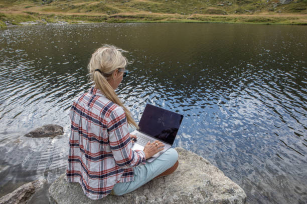 istock Woman sits on rock using laptop by the lake, remote working 1465525038