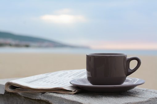 Ceramic cup of hot drink and newspaper on stone surface near sea in morning