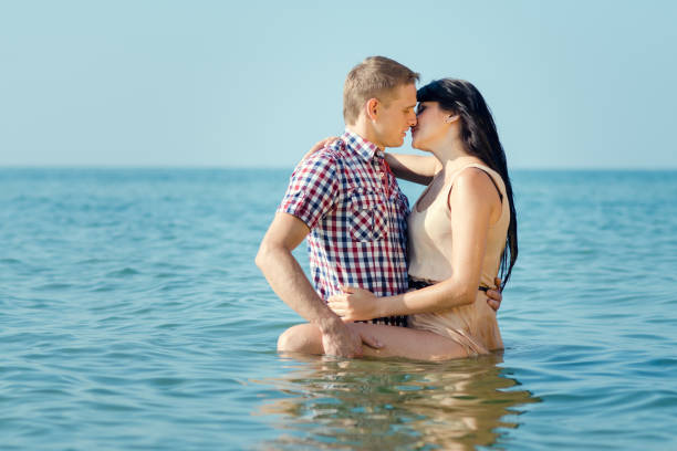 Swimming in clothes. Portrait of a beautiful man and woman wet in sea. Couple hugging and kissing in sea. Male kisses and hugs female standing on water with big waves ocean and enjoying a summer day.