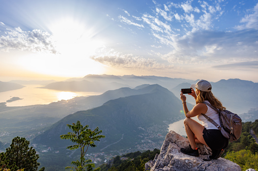 A happy contented young girl with a backpack takes a souvenir photo of the Montenegrin coastal cities, the Adriatic Sea and the bright sunset rays from the top of a high mountain