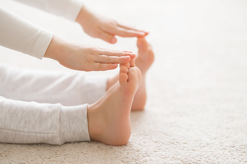 Little child in white sport clothes sitting on light carpet and doing exercise at room. Stretching arms to toes. Barefoot closeup. Active training time at home. Side view.