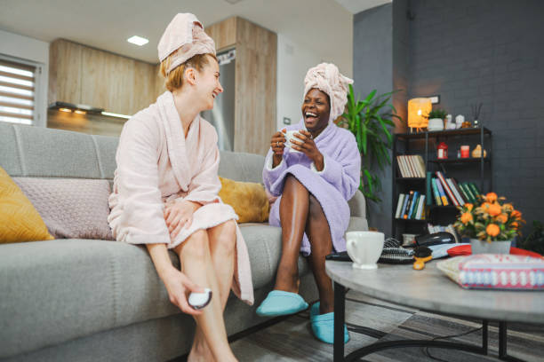 dos amigas multirraciales que tienen un relajante día de spa en casa - bathrobe women cup towel fotografías e imágenes de stock