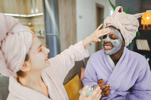Two multiracial female friends putting on face mask and having a relaxing spa day at home