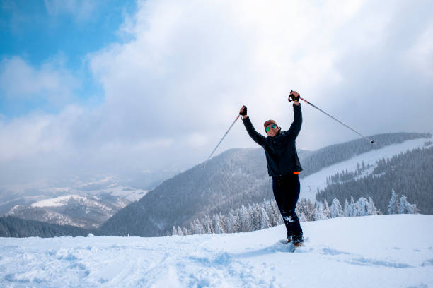 Young Man with hands up holding hiking poles celebrate success in cold winter day. Active Male in sports warm clothes happy on winter recreation. Copy space