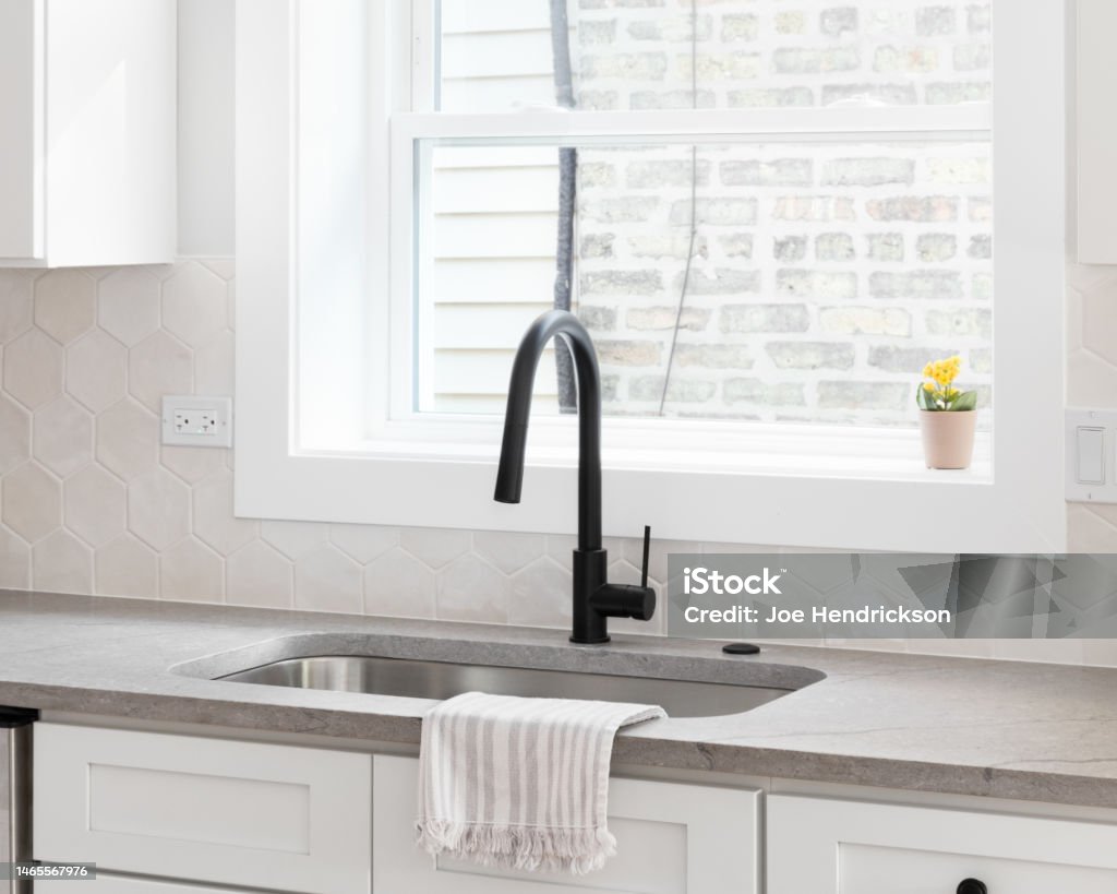 A cozy kitchen sink detail with a hexagon backsplash. A beautiful kitchen sink detail with white cabinets, marble countertops, and a hexagon tiled backsplash. Black Color Stock Photo