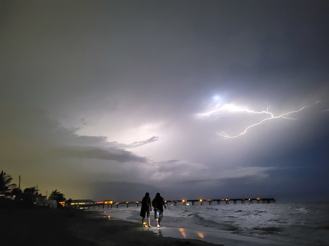 A couple walks along the night shore with a dock in the distance and a flash of lightning illuminating the sky