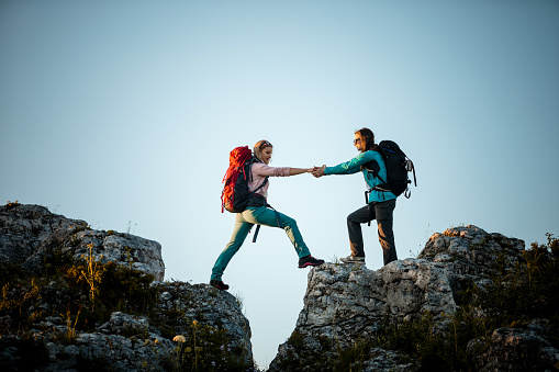 Side view of two young woman wearing sports clothes and backpacks helping her friend hiking the rocky hill at sunset.