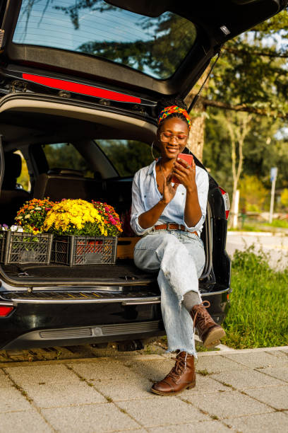 istock Young woman sitting in open car trunk, using smart phone 1465580296