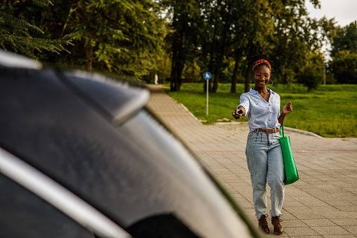 Selective focus shot of young woman walking up to her car on the parking lot, unlocking it with a car key, about to load in groceries in a reusable shopping bag in the car trunk.