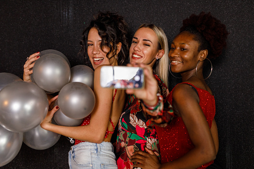 Selective focus shot of three joyful girlfriends standing against black background and taking selfies with smart phone during a New Year's eve party celebration.