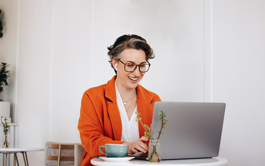 Mature businesswoman smiling cheerfully during a virtual meeting with a client in a cafe. Successful business woman having a video call on a laptop while working remotely.