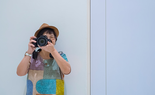 Front view of Asian female tourist photographing with digital camera while leaning against the wall inside of white room