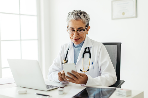 Female general practitioner using a smartphone at her desk. Mature doctor using smart medical apps to manage her patient schedules. Tech-savvy doctor working in her office.