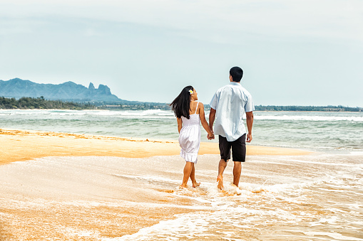 Photo of a happy Hawaiian couple walking in the surf along a remote beach, holding hands as they walk away from the camera
