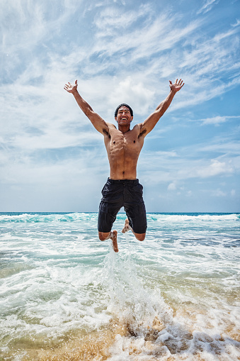 Photo of a young, muscular Hawaiian man in navy blue swim trunks, jumping for joy at the water's edge on a remote Hawaiian beach