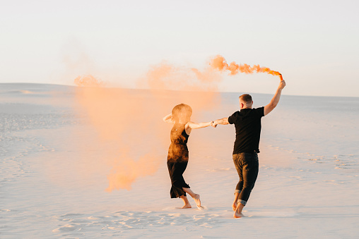 guy and a girl in black clothes hug and run on the white sand with orange smoke