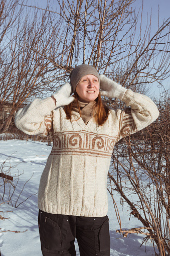 A happy woman in a beige patterned sweatshirt and mittens adjusts her hat against the background of a winter landscape