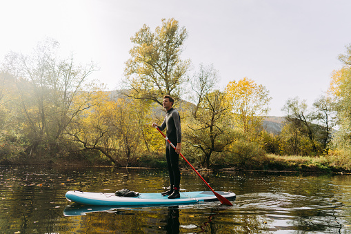 Photo of a young man stand-up paddle boarding on the  river; enjoying the beautiful, calm autumn afternoon, far from the hustle of the city.