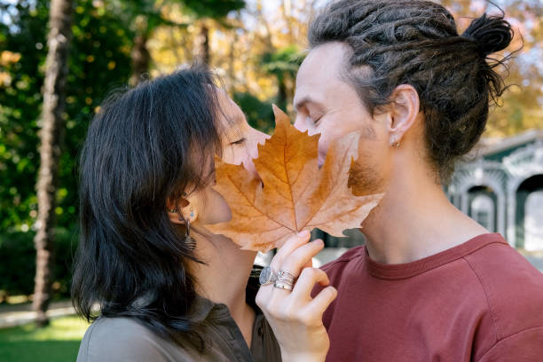 couple kisses and closes with maple yellow leaf in autumn park, romantic happy date. concept of love, relationship, privacy. - human face heterosexual couple women men imagens e fotografias de stock