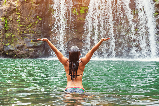 Rear-view photo of a young Hawaiian woman wading in a pool near a remote Hawaiian waterfall with arms outstretched skyward in joy