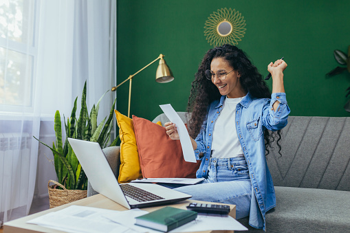 Happy hispanic woman at home doing paperwork and calculating household budget, sitting on couch in living room and using laptop at work, holding hand up victory and triumph gesture.