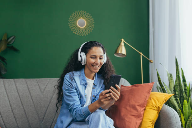 joven hispana sonriente en casa con auriculares y teléfono escuchando música en línea usando la aplicación de teléfono inteligente sentada en el sofá de la sala de estar y bailando - turkish ethnicity white black contemporary fotografías e imágenes de stock