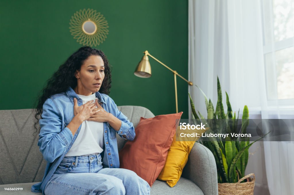 A Latin American asthmatic woman is experiencing an asthma attack. Holding his chest with his hands A young Latin American asthmatic woman is experiencing an asthma attack. He is sitting at home on the sofa, holding his chest with his hands, it is difficult to breathe, he needs medical help. Toughness Stock Photo