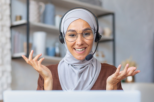 Joyful woman in hijab working from home remotely, Muslim woman talking on video call using headset and laptop, businesswoman working remotely at home.