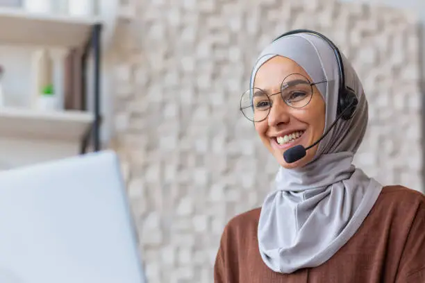 Photo of Joyful woman in hijab working from home remotely, Muslim woman talking on video call using headset and laptop, businesswoman working remotely at home