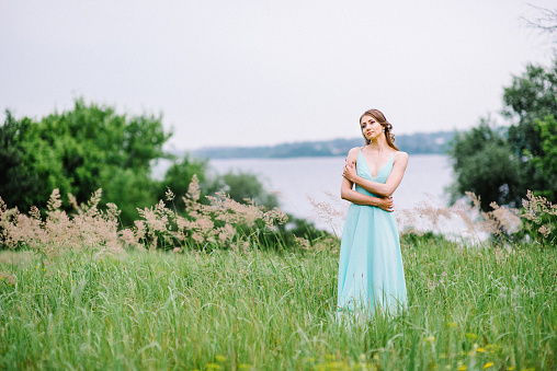 Romantic picture of young pretty model in dress sitting on rock with beautiful green field in background
