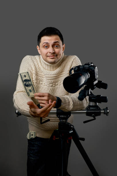 Professional photographer with digital camera on tripod holding hundred dollar bills on gray background