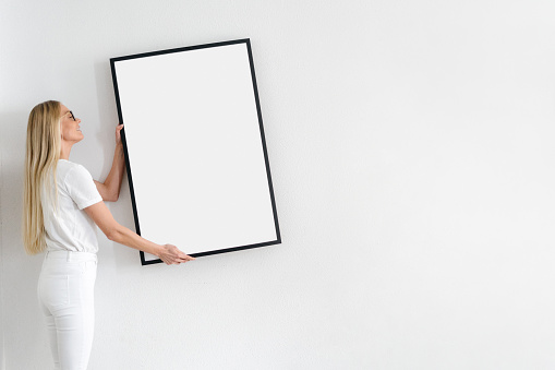 smiling female holding blank poster in frame and decorate wall on white background in living room, domestic interior concept