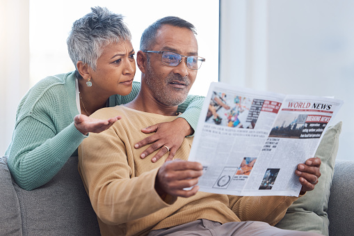 Senior couple, shocked and surprised by news while reading newspaper on sofa in living room at home together. Old man and woman check media article with fake news or bad crisis headline in retirement
