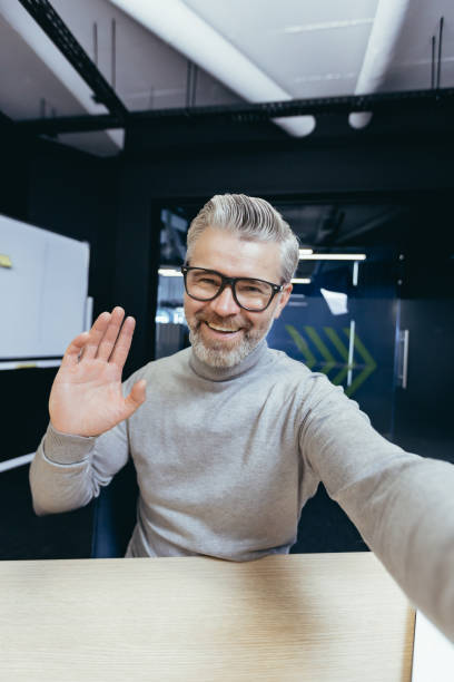 Mature man inside office talking on video call using app on phone, senior businessman having fun talking with friends and colleagues, taking selfie photo Mature man inside office talking on video call using app on phone, senior businessman having fun talking with friends and colleagues, taking selfie photo. man video call pov stock pictures, royalty-free photos & images