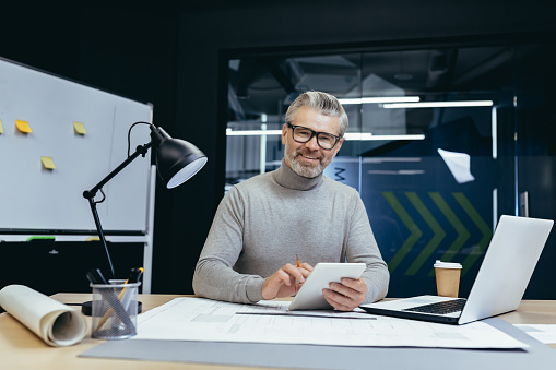A senior gray-haired man, architect, designer, engineer. Sitting in the office with a laptop and paperwork. He holds a notebook with a pencil in his hands. He looks at the camera and smiles.