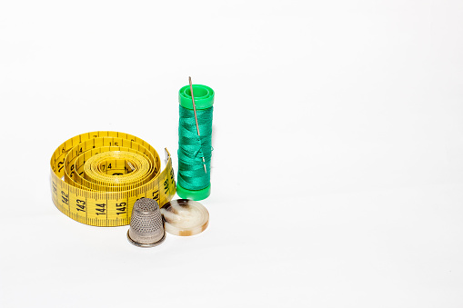 Spool of green thread with a pin pinned together with a yellow tape measure, a brown button and a metal thimble on a white background.