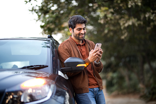 Smiling young man texting on mobile phone leaning on her car