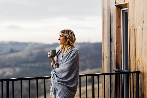 A pensive smiling Caucasian female covered with a blanket looking away while drinking tea outdoors.