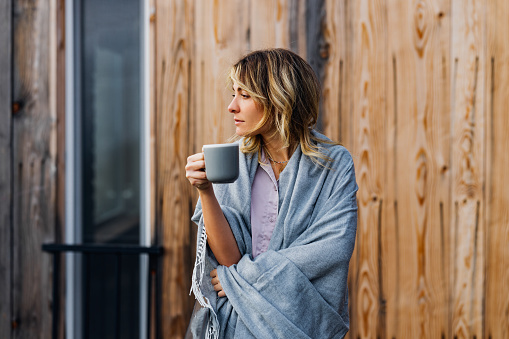 A pensive smiling Caucasian female wrapped in blanket looking away while drinking tea outdoors.