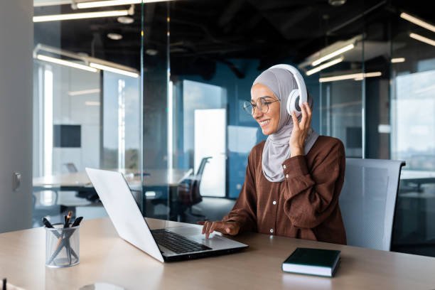 donna d'affari moderna di successo che lavora all'interno dell'ufficio con laptop, donna musulmana che indossa hijab e cuffie che ascolta audiolibri e podcast sul posto di lavoro, donna soddisfatta e di successo - the thinker audio foto e immagini stock