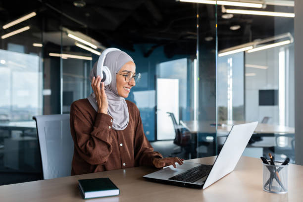 donna d'affari moderna di successo che lavora all'interno dell'ufficio con laptop, donna musulmana che indossa hijab e cuffie che ascolta audiolibri e podcast sul posto di lavoro, donna soddisfatta e di successo - the thinker audio foto e immagini stock