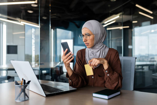 donna sconvolta e delusa in hijab all'interno dell'ufficio cercando di fare acquisti nel negozio online, donna d'affari che tiene lo smartphone e la carta di credito bancaria, ha ricevuto l'errore di trasferimento di denaro - poor communication foto e immagini stock