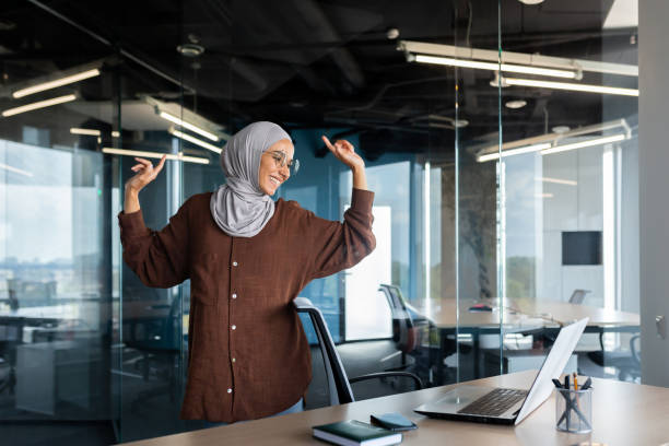 Joyful and successful businesswoman dancing at workplace, Muslim woman in hijab celebrating victory and successful achievement at work, having a good mood inside the office, Arab at work with laptop. mujer de negocios alegre y exitosa bailando en el lugar de trabajo, mujer musulmana con hijab celebrando la victoria y el logro exitoso en el trabajo, teniendo buen humor dentro de la oficina, árabe en el trabajo con computadora portátil