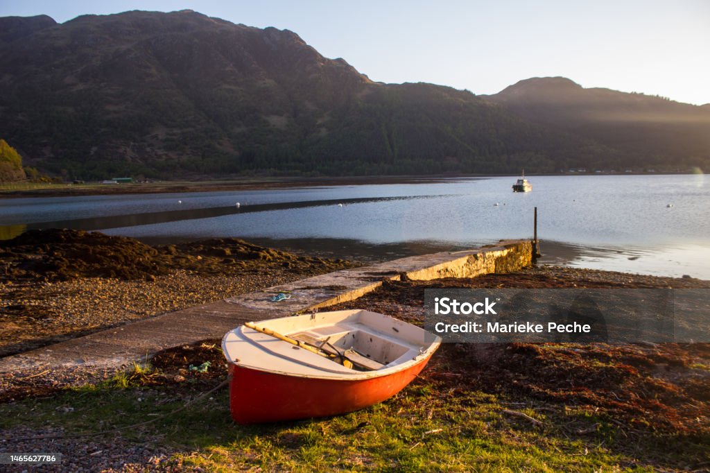 Small red rowboat, on the shores of Loch Duich in the golden light of late afternoon Small red rowboat, beached on the shores of Loch Duich, Scotland, in the late afternoon. Loch Duich is a sea loch situated on the western coast of the Scottish Highlands. It is famous for its connection to local legend regarding Selkies. Nautical Vessel Stock Photo